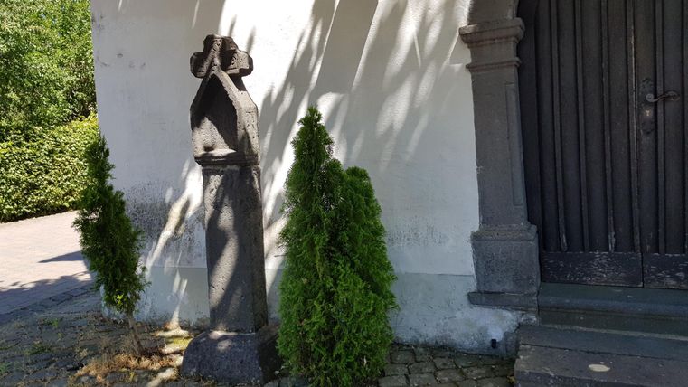 An old statue stands against the wall of a building, surrounded by two small bushes. The access to the door is made of stone and well-maintained.