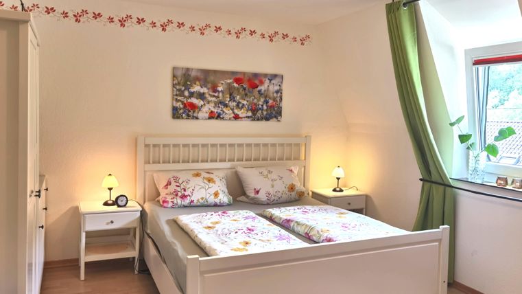 Ein gemütliches Schlafzimmer mit einem weißen Bett und floralen Bettwäsche. Das Zimmer hat große Fenster und eine freundliche Atmosphäre.