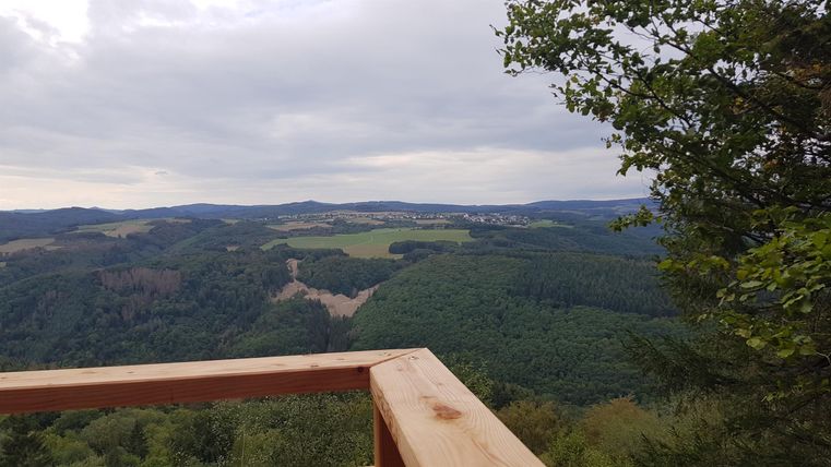 Eine Aussicht auf eine grüne Hügellandschaft unter einem bewölkten Himmel. Im Vordergrund ist ein Holzgeländer sichtbar.