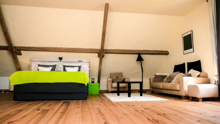 A modern bedroom with a double bed and a cozy living area. The room has a wooden floor and light walls with large windows.