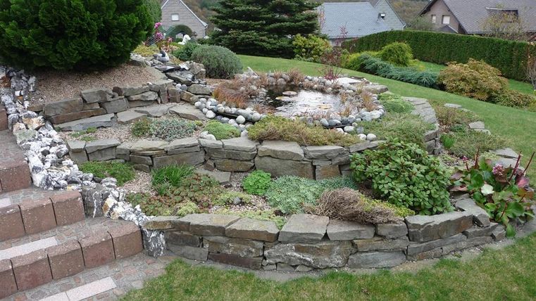 A beautifully designed garden with various plants and stones. In the background, some houses and a well-maintained lawn can be seen.