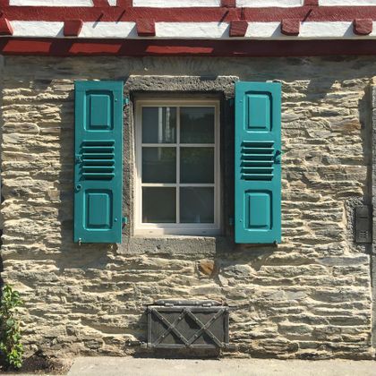 Ein Fenster mit grünen Fensterläden in einer Steinfassade. Der obere Teil des Hauses hat rote Holzbalken.