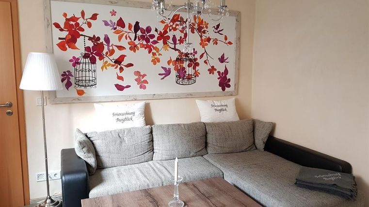 A cozy living room with a gray sofa and a wooden coffee table. A colorful wall tapestry with floral designs and cages hangs on the wall.