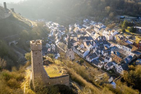 An impressive ruin on a hill overlooks a picturesque village. The houses are surrounded by green forests and gentle hills.