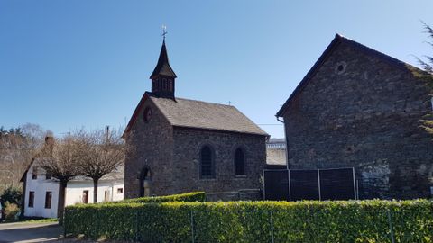 Eine alte Kirche mit einem hohen Turm steht neben einem anderen Gebäude. Der Himmel ist klar und blau, umgeben von grüner Bepflanzung.