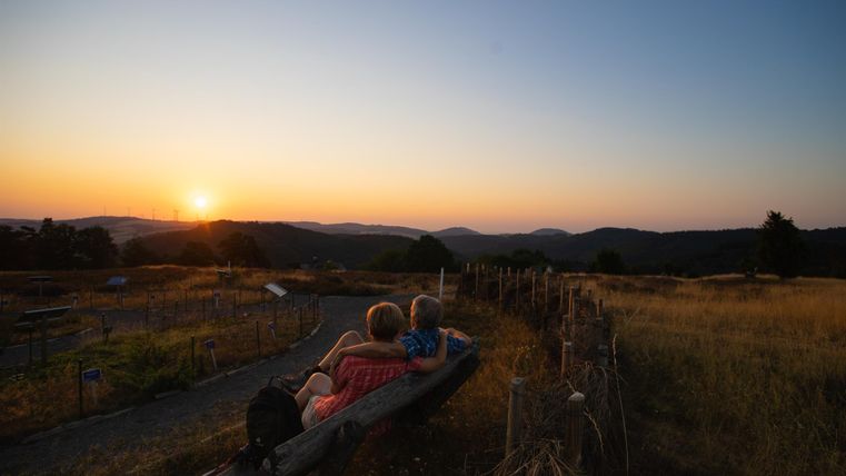 Ein Paar sitzt auf einer Bank und beobachtet den Sonnenuntergang über den Bergen. Die Landschaft ist sanft und friedlich, mit goldenen Wiesen und einem klaren Abendhimmel.