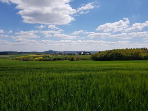 Eine weite grüne Landschaft mit sanften Hügeln unter einem blauen Himmel. Wolken ziehen vorbei und im Hintergrund ist ein kleines Dorf zu sehen.