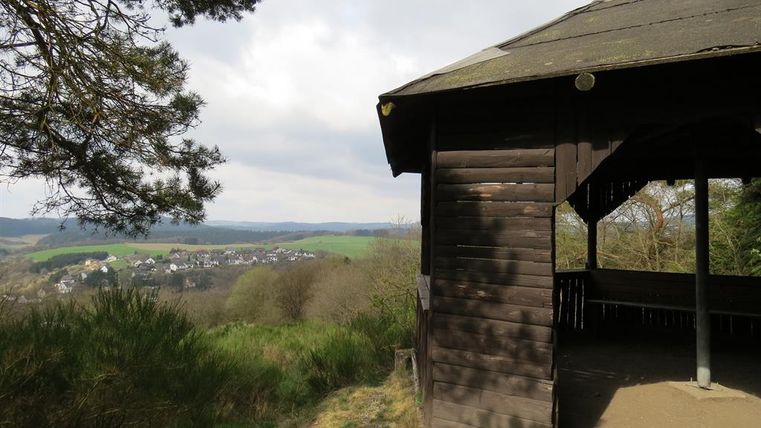 A wooden gazebo stands on a hill overlooking a green valley. In the background, small houses and a wooded landscape can be seen.