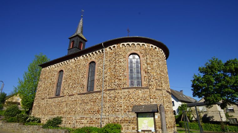 Eine historische Kirche aus Steinen mit einem markanten Kirchturm. Umgeben von Grün und blauem Himmel.