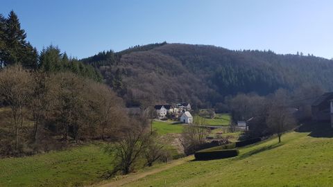 Eine ruhige Landschaft mit sanften Hügeln und grünen Wiesen. Im Hintergrund sind einige Häuser und Bäume sichtbar.