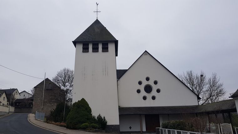 A modern church with a tall tower and round windows. It stands in a rural setting with trees and a small path.