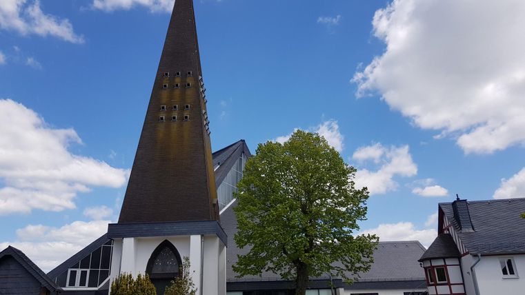 Eine moderne Kirche mit einem hohen, spitzen Turm und großen Fenstern. Im Vordergrund steht ein grüner Baum vor einem blauen Himmel mit wenigen Wolken.