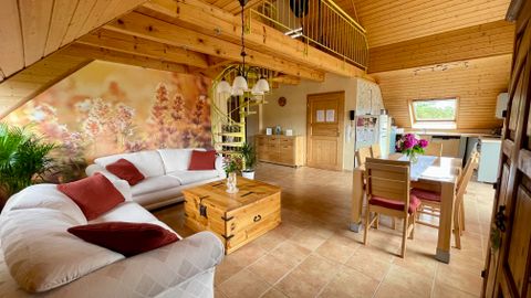 Ein gemütlicher Wohnraum mit einer weißen Couch, einem Holztisch und Stühlen. Die Wände sind mit einem Blumenmuster dekoriert, und es gibt viel Tageslicht.