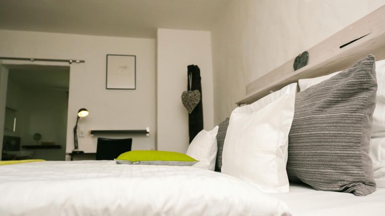 A modern bedroom with a large bed and white and gray pillows. In the background, there is a desk and decorative elements.