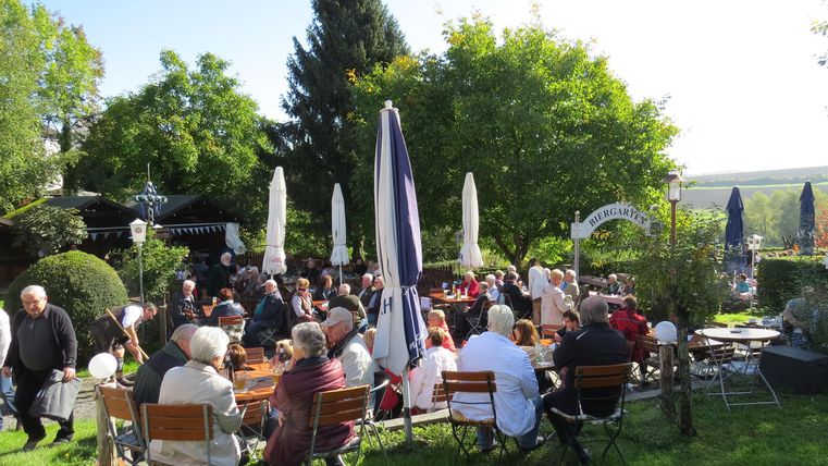 Eine lebhafte Gartenveranstaltung mit vielen Gästen, die an Tischen sitzen. Umgeben von Bäumen und Sonnenschirmen, genießt die Gruppe den schönen Tag.