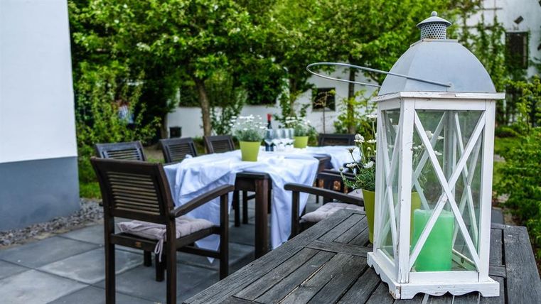 An inviting garden with a table and chairs, decorated with a white lantern. In the background, green plants and a set table are visible.