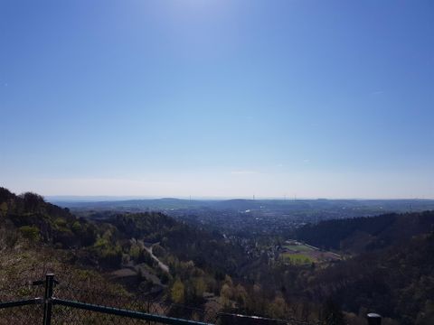 Eine weite Aussicht auf das Tal unter einem klaren blauen Himmel. Grüne Hügel und Bäume prägen die Landschaft im Vordergrund.