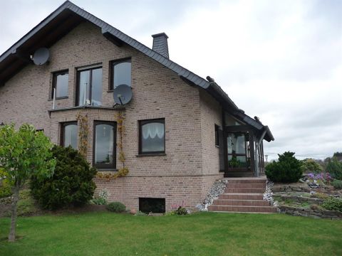 Ein zweigeschossiges Haus aus Ziegel mit großen Fenstern und einem modernen Eingang. Der Garten ist gepflegt und mit grünen Bäumen und Sträuchern gestaltet.