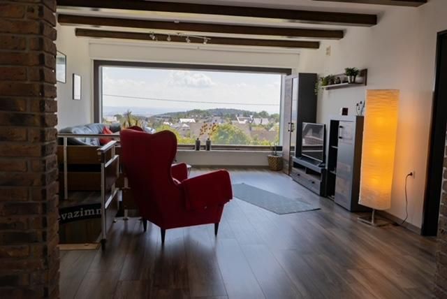 Ein gemütliches Wohnzimmer mit einem roten Sessel und Blick auf eine hügelige Landschaft. Große Fenster sorgen für viel Tageslicht.