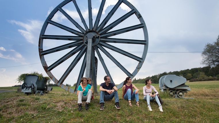 Eine Gruppe von vier Personen sitzt entspannt auf einer Wiese vor einem großen, alten Rad. Im Hintergrund sind landwirtschaftliche Geräte und ein bewölkter Himmel sichtbar.