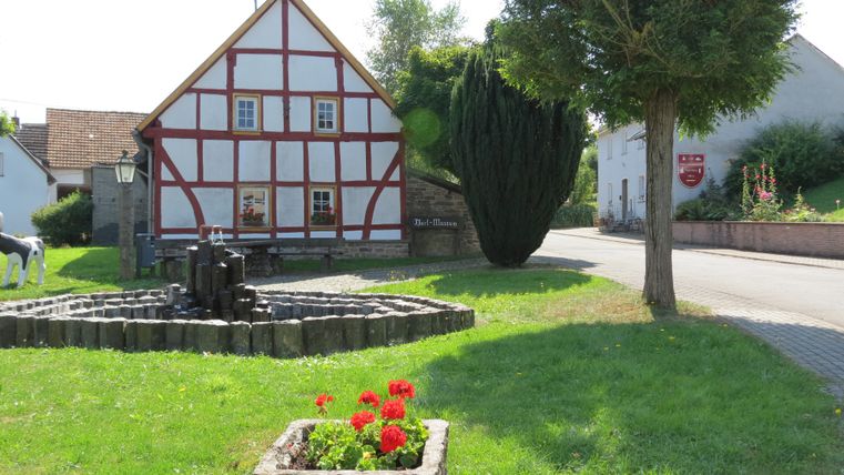 Fachwerkhaus mit Brunnen und Blumenbeet im Vordergrund.