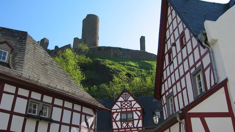 A picturesque view of historic half-timbered houses and a castle ruin on a hill. The buildings are designed with red and white beams, surrounded by green countryside.