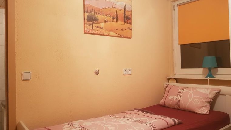 A simple bedroom with a single bed and a colored pillow. A painting hangs on the wall and the window has an orange blind.