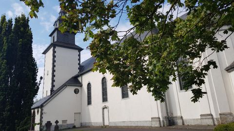 Eine weiße Kirche mit einem hohen Turm steht unter einem klaren Himmel. Umgeben von grünen Bäumen wirkt sie einladend und ruhig.