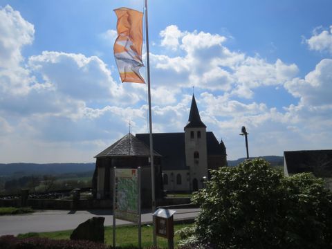 Ein malerisches Dorf mit einer Kirche im Hintergrund und einer Fahne, die im Wind weht. Der Himmel ist klar mit einigen Wolken, was eine ruhige Atmosphäre schafft.