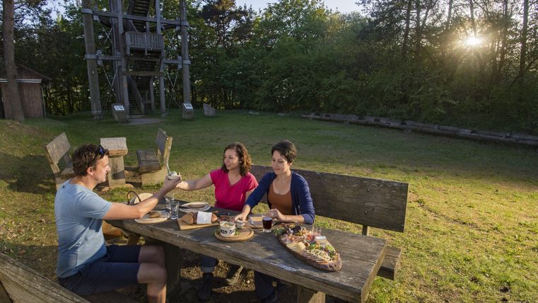 Three people are sitting at a wooden table outdoors, enjoying a meal. A lookout tower can be seen in the background.