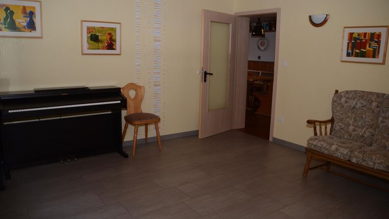 Ein heller Raum mit einem Klavier, einem Stuhl und einer Couch. An den Wänden hängen einige Bilder.
