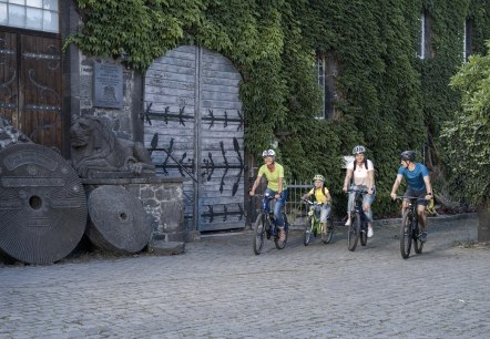 Eine Familie mit Helmen f&auml;hrt auf Fahrr&auml;dern an einem efeubewachsenen Geb&auml;ude mit einer L&ouml;wenstatue vorbei. Der Boden ist mit Kopfsteinpflaster bedeckt., &copy; Klaus-Peter Kappest/Vulkanregion Laacher See