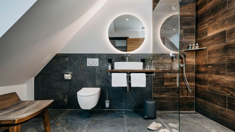 Ein modernes Badezimmer mit Holz- und Naturstein-Elementen. Es verfügt über eine Dusche, ein Waschbecken mit runder Spiegel und einen großen Fenster.
