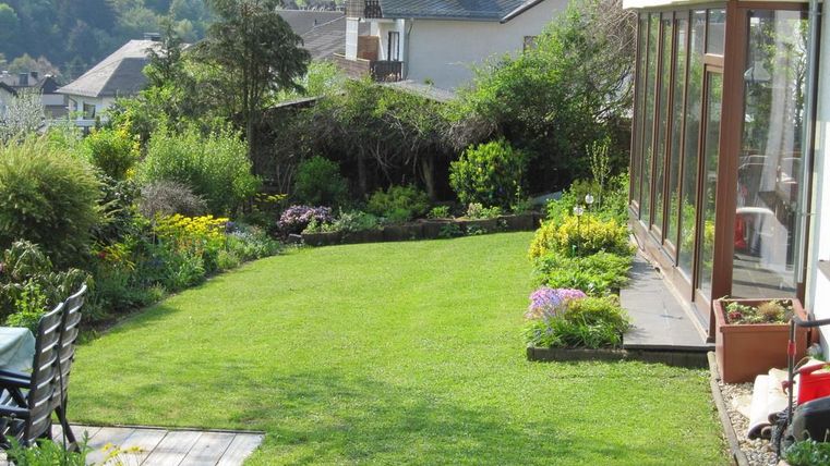Ein schöner Garten mit frischem, grünem Rasen und bunten Blumen. Im Hintergrund sind Häuser und Bäume zu sehen.