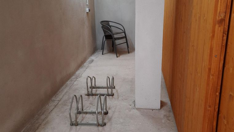A narrow hallway with walls in neutral shades. On the floor, there are two metal frames and a chair is placed against the wall.