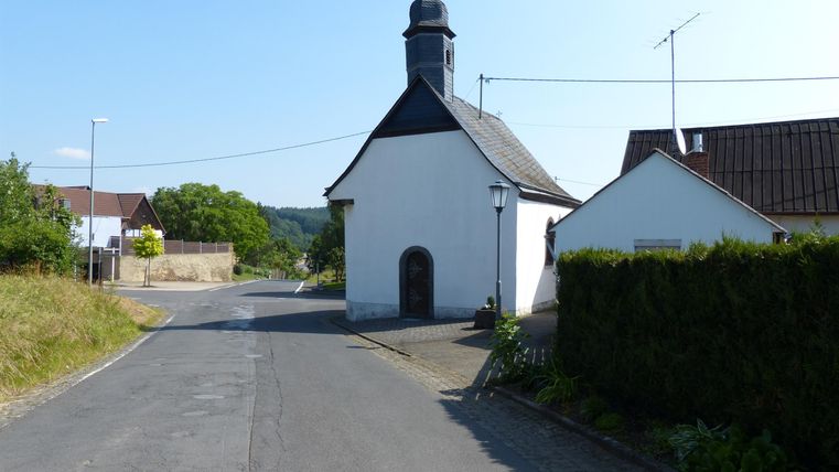 Eine kleine, weiße Kapelle steht an einer Straße in einer ländlichen Gegend. Der Himmel ist klar und blau, umgeben von Bäumen und Häusern.