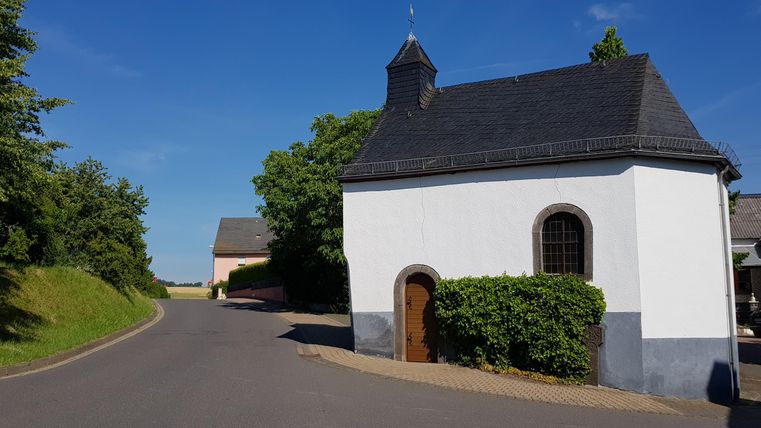Eine kleine, weiße Kapelle mit einem dunklen Dach steht an einer kurvigen Straße. Umgeben von Bäumen und grünem Gelände strahlt die Szene Ruhe und Idylle aus.