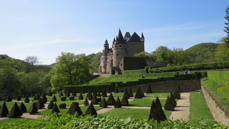An impressive castle surrounded by well-kept gardens and green hills. The sky is clear and blue.