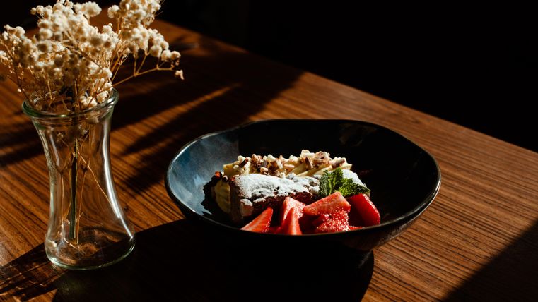 Ein schwarzes Schüssel mit frischem Obst und einer säuerlichen Creme steht auf einem Holztisch. Im Hintergrund ist eine Glasvase mit getrockneten Blumen zu sehen.