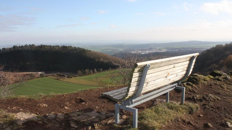 A wooden bench is sitting on a hill overlooking the landscape. In the background, fields and a clear sky can be seen.