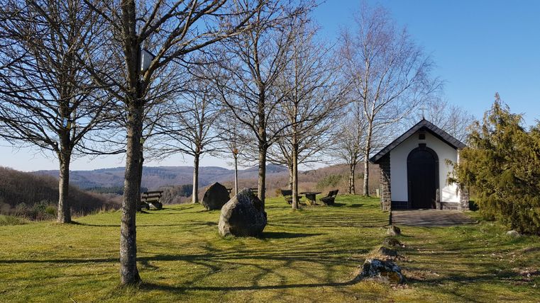 Eine ruhige Landschaft mit kahlen Bäumen und einer kleinen Kapelle. Im Hintergrund sind sanfte Hügel und ein klarer blauer Himmel zu sehen.