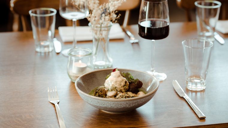 Ein eleganter Tisch mit einem köstlichen Gericht in einer Schüssel, umgeben von Gläsern und Besteck. Ein Glas Rotwein steht ebenfalls auf dem Tisch.