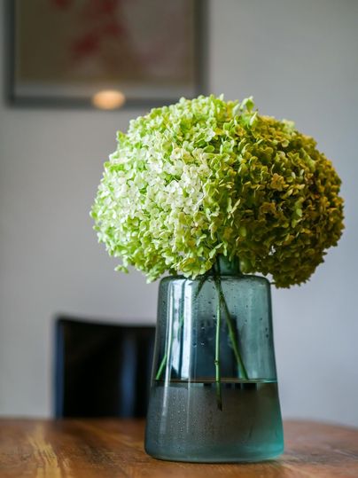 A large green floral ball in a transparent vase is placed on a wooden table. The background is blurry and shows a dark chair.