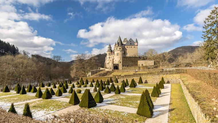 Eine majestätische Burg umgeben von einer gepflegten Gartenlandschaft. Der Himmel ist klar und Wolken ziehen sanft vorbei.