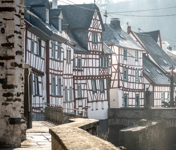 Fachwerkh&auml;user in Monreal, Deutschland, mit roten Balken und wei&szlig;en W&auml;nden. Eine Br&uuml;cke f&uuml;hrt &uuml;ber einen kleinen Fluss, im Hintergrund bewaldete H&uuml;gel., &copy; Eifel Tourismus GmbH, D. Ketz