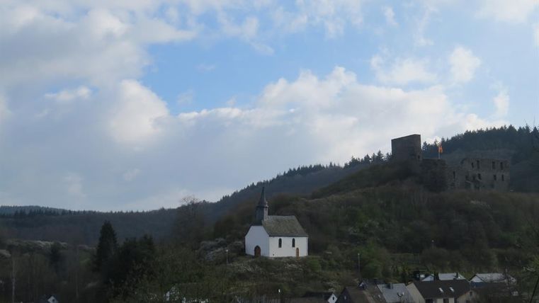 A small white church stands on a hill, surrounded by trees. In the background, the ruins of a castle are visible under a cloudy sky.