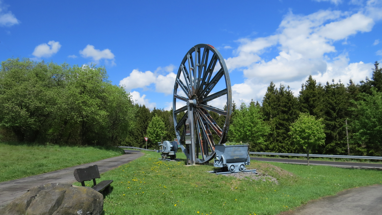 A large water wheel stands in a green landscape. In the background, trees and a blue sky with a few clouds are visible.
