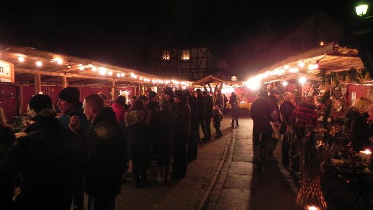 Ein geschäftiger Weihnachtsmarkt bei Nacht, beleuchtet von warmen Lichtern. Menschen genießen das festliche Ambiente und die Stände mit Handwerkskunst und Leckereien.