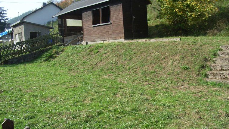 Ein kleines Holzhaus steht auf einer grünen Wiese. Im Hintergrund sind Bäume und ein weiterer Wohnort zu sehen.