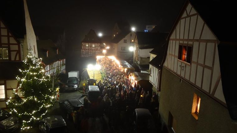 Eine festliche Szenerie bei Nacht mit einer großen Menschenmenge und einem beleuchteten Weihnachtsbaum. Im Hintergrund sind traditionelle Fachwerkhäuser zu sehen, die die Atmosphäre ergänzen.